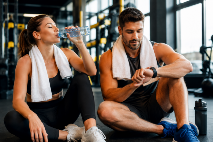 Sportlerin und Sportler trinken Wasser nach dem Training in einem modernen Fitnesspark, während sie ihre Trainingsdaten auf einer Smartwatch analysieren.